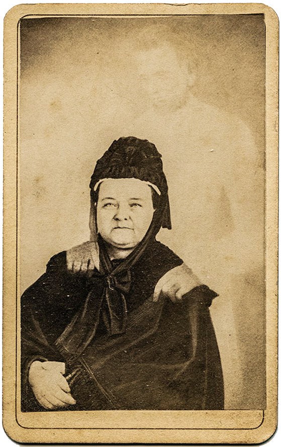 William Mumler's 'spirit photograph' of Mary Todd Lincoln, c.1872 — a seated woman in dark mourning dress with the pale, ghostly figure of a man (purportedly Abraham Lincoln) standing behind her, hands resting on her shoulders.