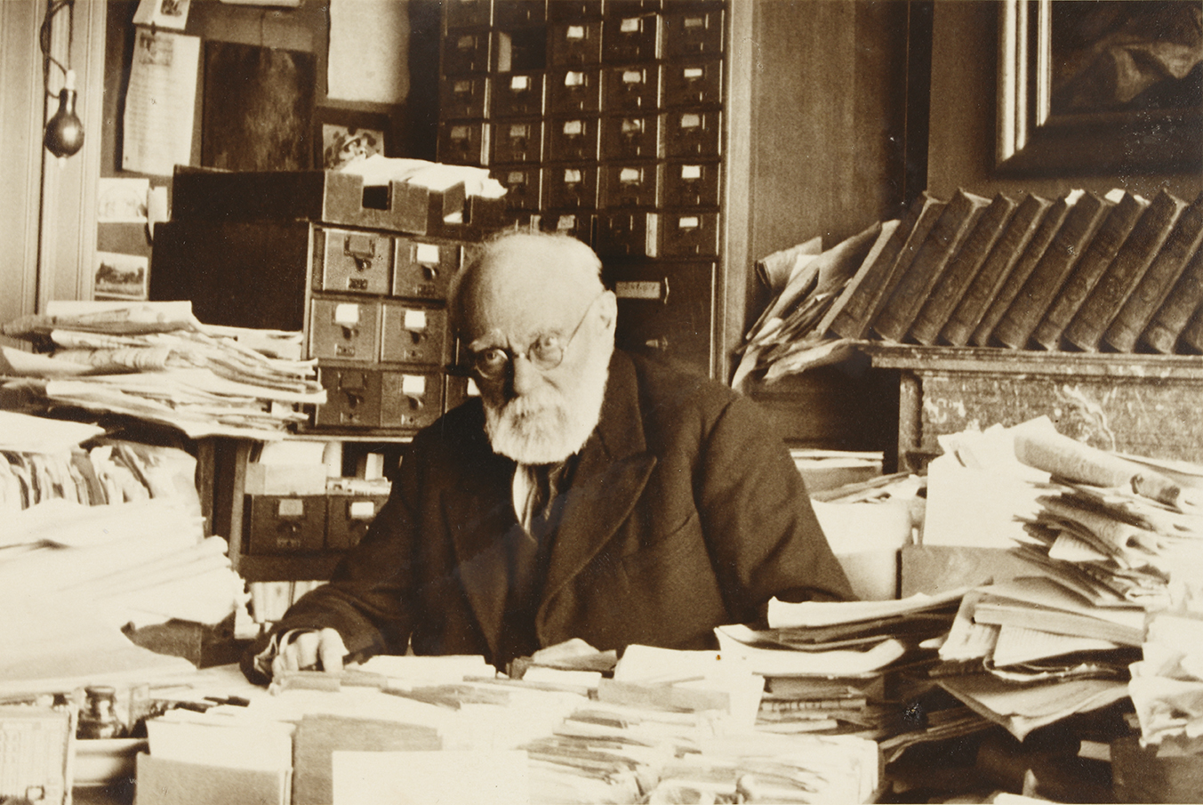 A 1937 photograph of Paul Otlet at his desk at the Mundaneum, surrounded by papers, drawers, and a card catalogue.