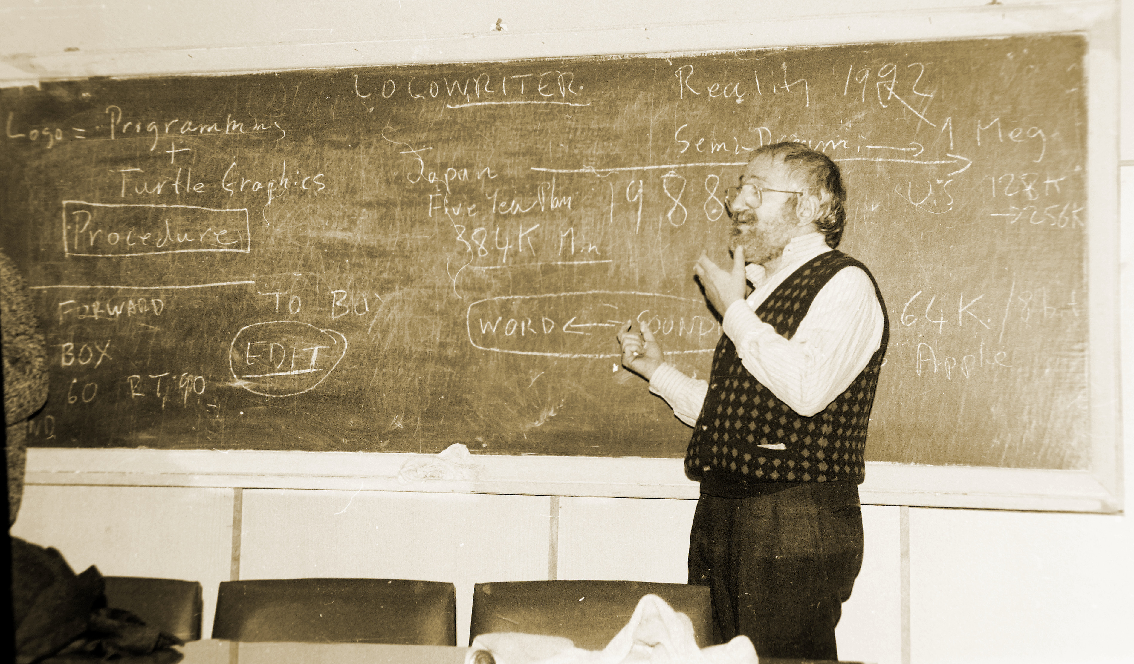 Seymour Papert standing at a blackboard covered in Logo syntax — FORWARD, BOX, RT 90, LogoWriter, Apple memory specs — addressing an audience in Moscow, 1987. Sepia-toned archival photograph.